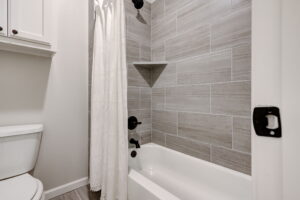 Bathroom with a white bathtub and shower, brown tiled walls, and dark fixtures. A white curtain hangs to the side, and a white toilet is partially visible.