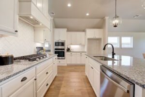 A modern kitchen with white cabinets, granite countertops, a central island with a sink, stainless steel oven and microwave, and a pendant light.