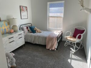 A cozy bedroom with a single bed, dresser, lamp, and decorative items. A window lets in natural light. The room features a white chair, plush toys, and floral wall art.