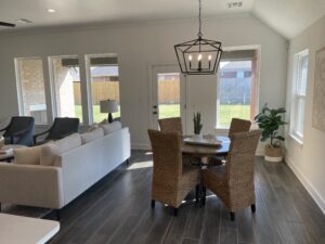 A modern living and dining area with gray tile flooring, wicker chairs around a round table, a white sofa, and large windows allowing natural light. A potted plant is by the window.