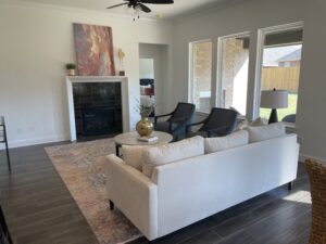 A modern living room with a white sofa, two black chairs, a round coffee table, and a fireplace. Large windows allow natural light in, and a ceiling fan is on the white ceiling.