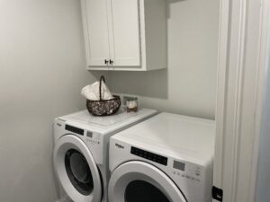 A laundry room features a front-loading washer and dryer set with a basket of folded towels on top and a wall cabinet above.