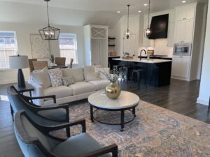 Open-plan living area with a beige sofa, patterned rug, and round coffee table. Kitchen in background features a black island and modern lighting fixtures.