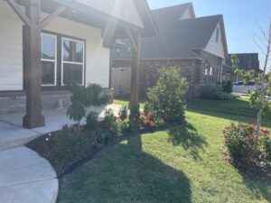 A front yard with a small porch, green grass, shrubs, and vibrant red flowers, next to a residential house.